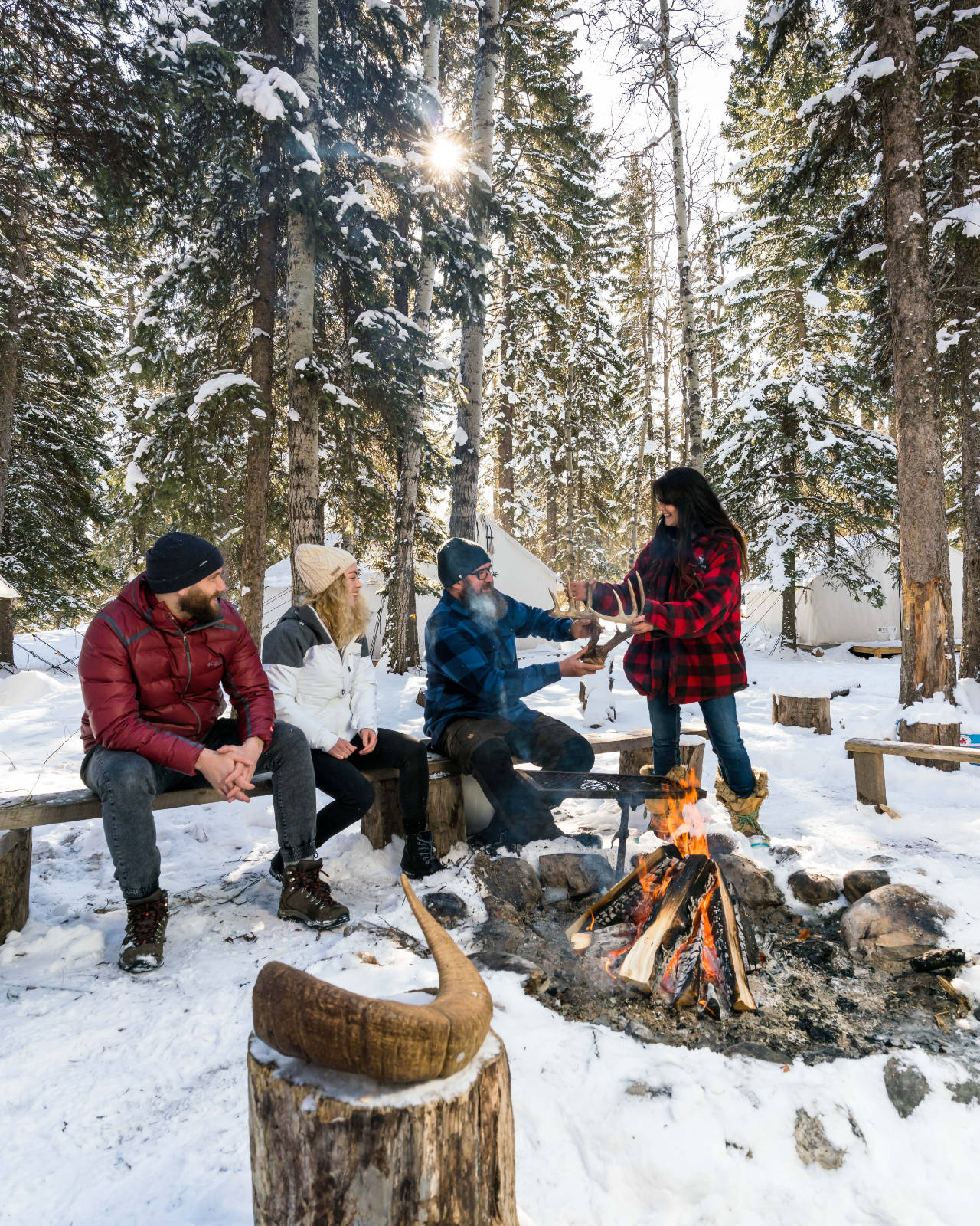A number of people sit on benches in a snowy tree filled scene around a fire.