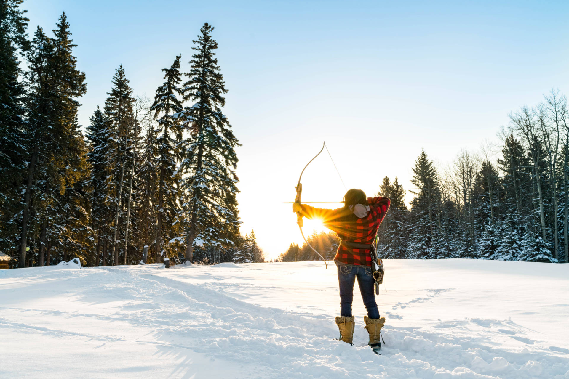 A bow and arrow are drawn back by a person in a backlit clearing surrounded by snow and trees.